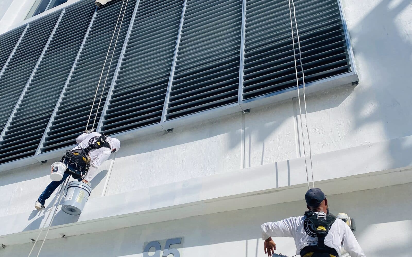 Two Basile crew members at Miami Arts Charter School, one suspended on rope access with a paint bucket beside the louvred facade