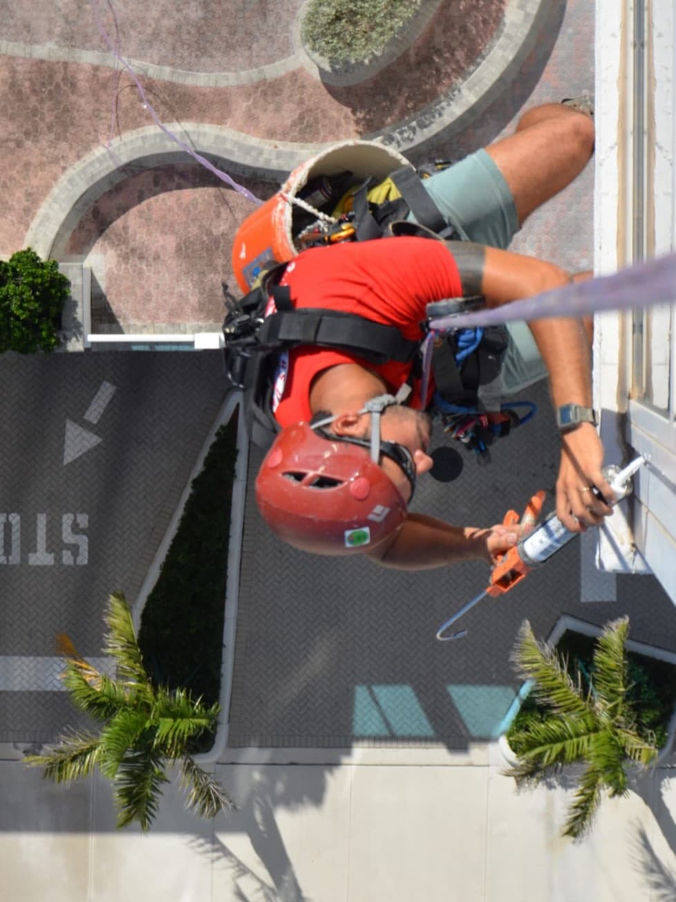 View straight down from a high-rise as a rope-access technician applies sealant, with palm trees far below